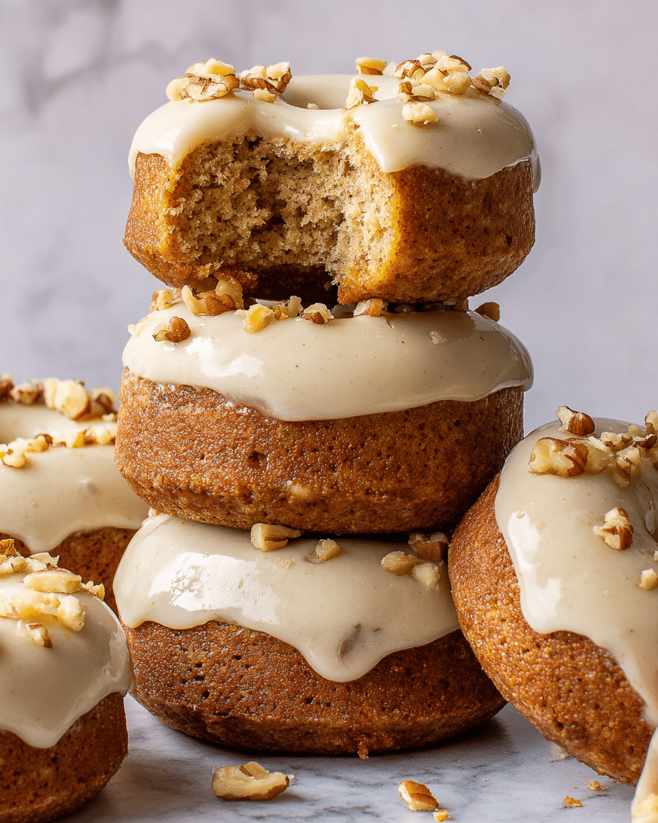 A stack of four thick, round carrot cake donuts is shown against a white marbled texture. Each donut has a light brown, moist texture and is covered with a glossy, creamy white frosting that drips slightly over the edges. Small pieces of chopped walnuts are sprinkled on top of the frosting and around the base of the donuts. The top donut has a bite taken out, revealing a dense and crumbly interior. The donuts are arranged in a way that the top bitten donut is balanced on the second one, with other donuts surrounding the main stack. photo taken with an iphone --ar 4:5 --v 7