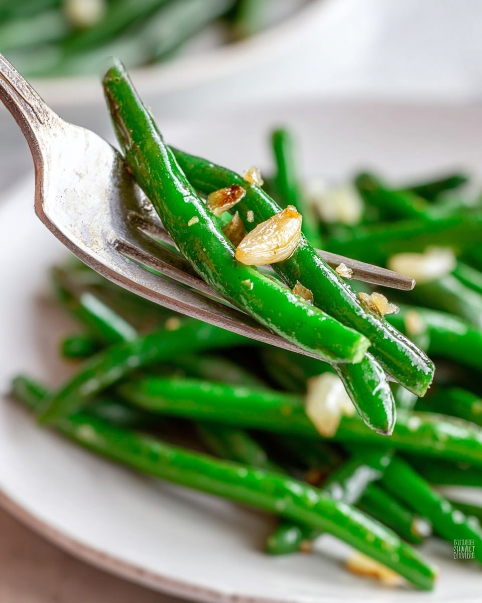 A close-up image shows a silver fork holding a small pile of shiny, cooked green beans with a few small pieces of golden-brown garlic on top. The green beans are bright green and look tender but firm. In the blurred background, a white plate with more green beans and garlic sits on a white marbled surface. The focus is sharp on the green beans on the fork, with soft light highlighting their glossy texture. photo taken with an iphone --ar 4:5 --v 7