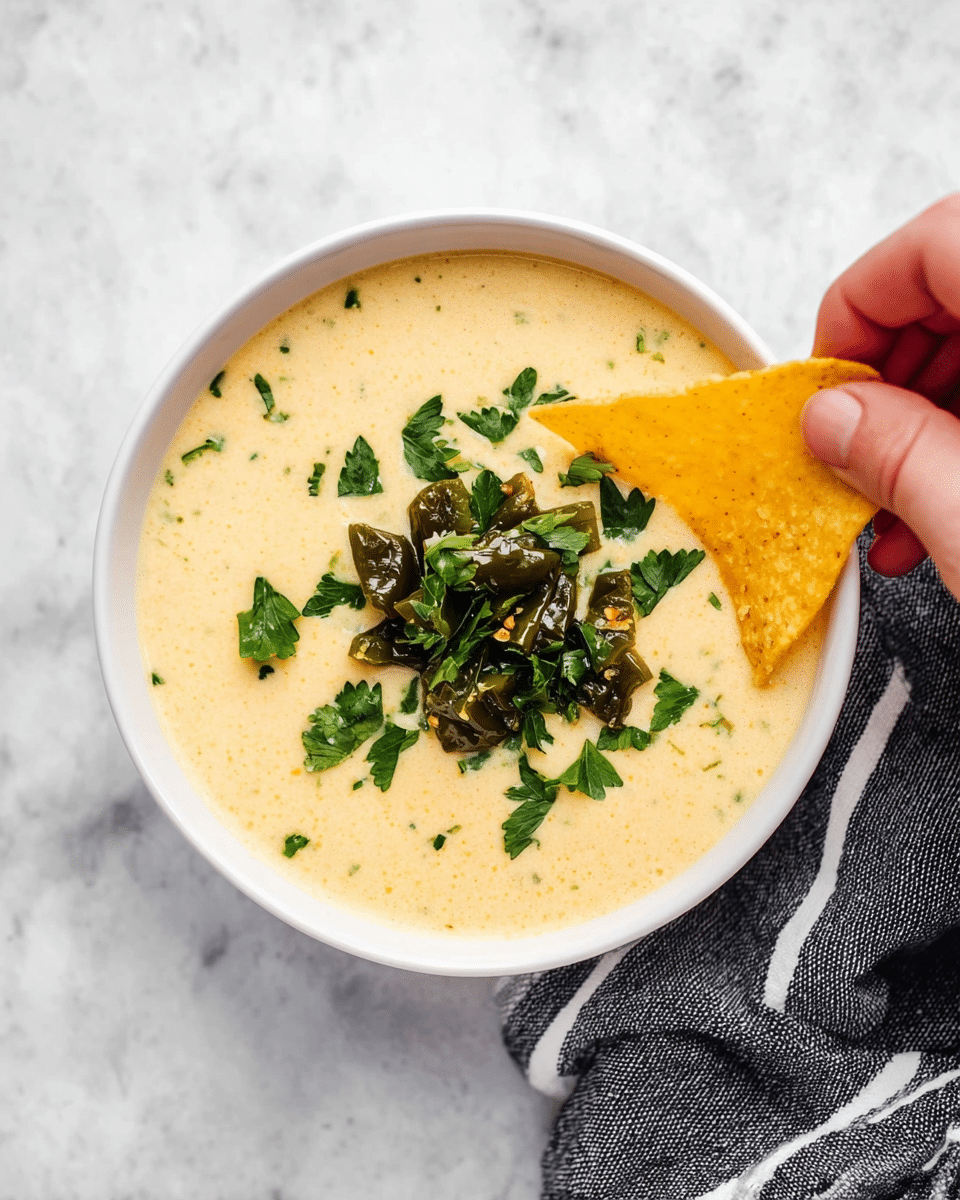 A white bowl filled with smooth, creamy cheese soup in a light yellow color, topped with a small pile of dark green roasted peppers and scattered bright green fresh parsley leaves at the center. A woman's hand is dipping a triangular yellow tortilla chip into the soup from the upper right side. The bowl is placed on a white marbled surface with part of a black-and-white striped cloth visible at the bottom right corner. Photo taken with an iphone --ar 4:5 --v 7