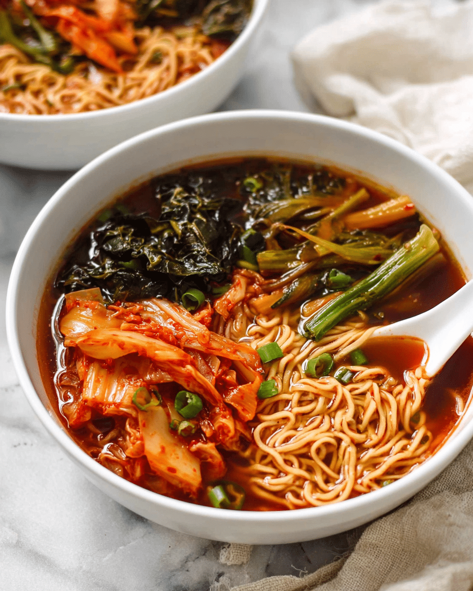 The image shows a white bowl filled with a colorful noodle soup placed on a white marbled surface with a cream cloth nearby. The bowl contains light brown noodles layered at the bottom, soaked in a deep reddish-brown broth. On top, there are several pieces of cooked kimchi with reddish-orange and pale beige hues and some cooked green leafy vegetables with dark green, slightly wilted texture. There are also long pieces of cooked carrot sticks in bright orange and green scallions, both laying on the noodles. A white ceramic soup spoon rests inside the bowl on the right side, partly covered in broth with some chopped green onions floating on it. The close-up view shows a second similar bowl blurred in the background. photo taken with an iphone --ar 4:5 --v 7