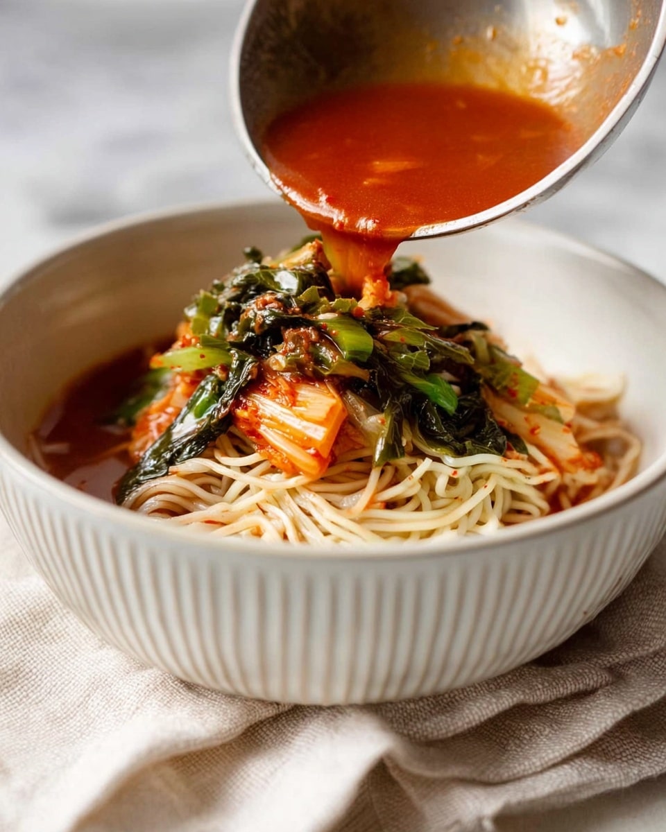 A close-up view of a bowl of light-colored thin noodles at the bottom, topped with chopped green leafy vegetables and reddish-orange kimchi. A ladle pours bright red, slightly thick soup with pieces of vegetables and herbs over the noodles, creating a mix of vibrant colors and textures. The bowl is white with vertical ridges and sits on a soft, off-white cloth over a white marbled surface. photo taken with an iphone --ar 4:5 --v 7