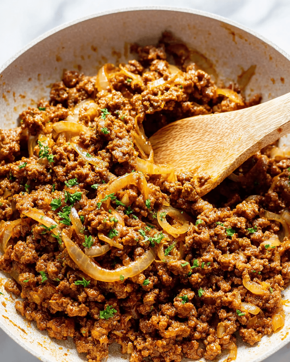 The image shows a close-up of a white bowl filled with browned ground meat mixed with cooked sliced onions. The meat is a rich brown color with a slightly crumbly texture, and the onions are translucent light yellow, scattered throughout the mix. Small bits of fresh chopped green herbs are sprinkled on top for contrast. A light wooden spoon is partially inserted into the mixture, with a small curl of onion caught in it. The background has a white marbled texture. Steam rises gently from the warm food, adding a fresh, just-cooked feeling. Photo taken with an iphone --ar 4:5 --v 7