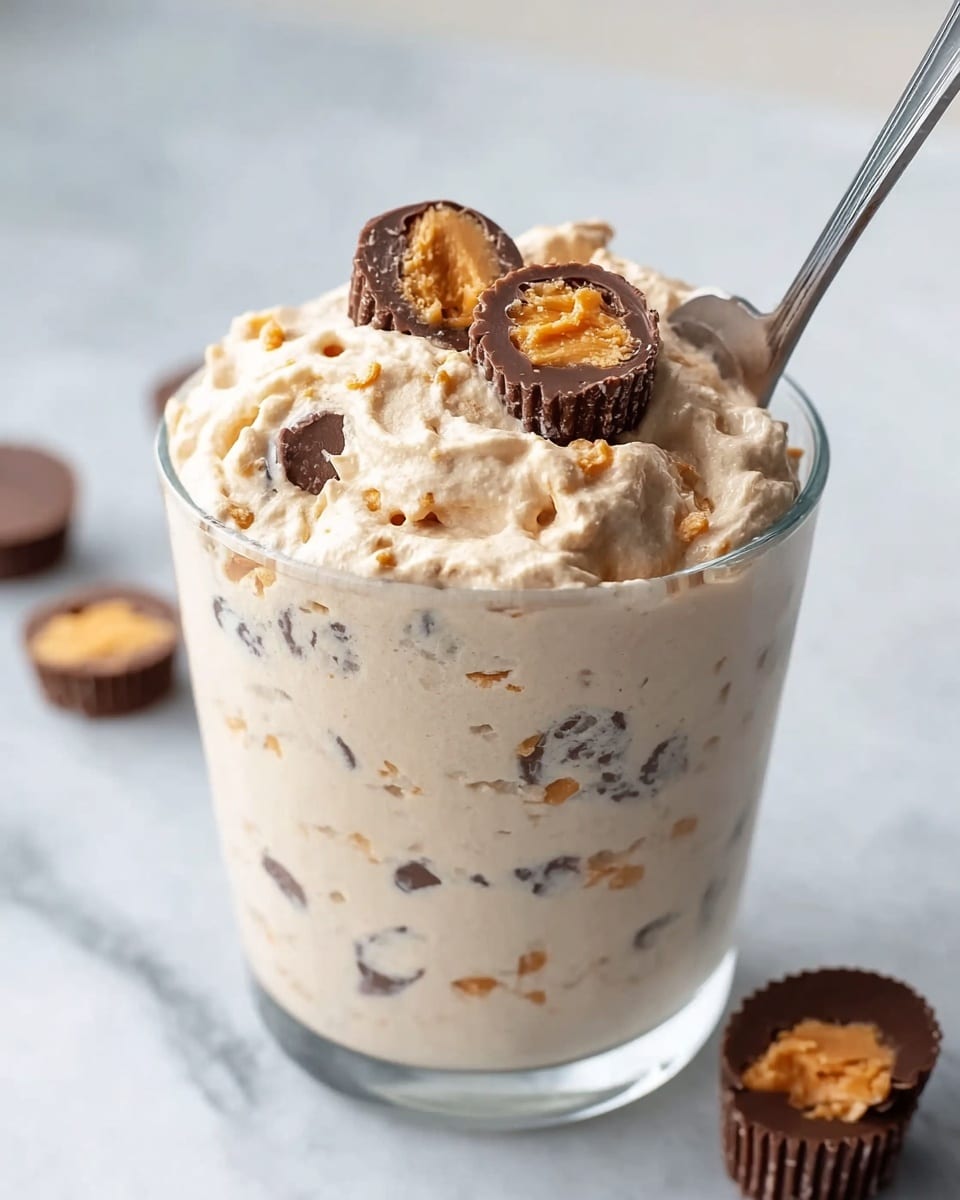 A clear glass filled with a creamy, light beige mixture that looks thick and fluffy, with visible small chunks of chocolate and peanut butter throughout. On top, several mini chocolate peanut butter cups are placed, some halved to show the creamy orange filling inside. A shiny silver spoon is inserted into the mixture from the right side, with the creamy texture slightly mounded above the rim of the glass. The glass sits on a white marbled surface, and a few more mini peanut butter cups are blurred in the background. photo taken with an iphone --ar 4:5 --v 7