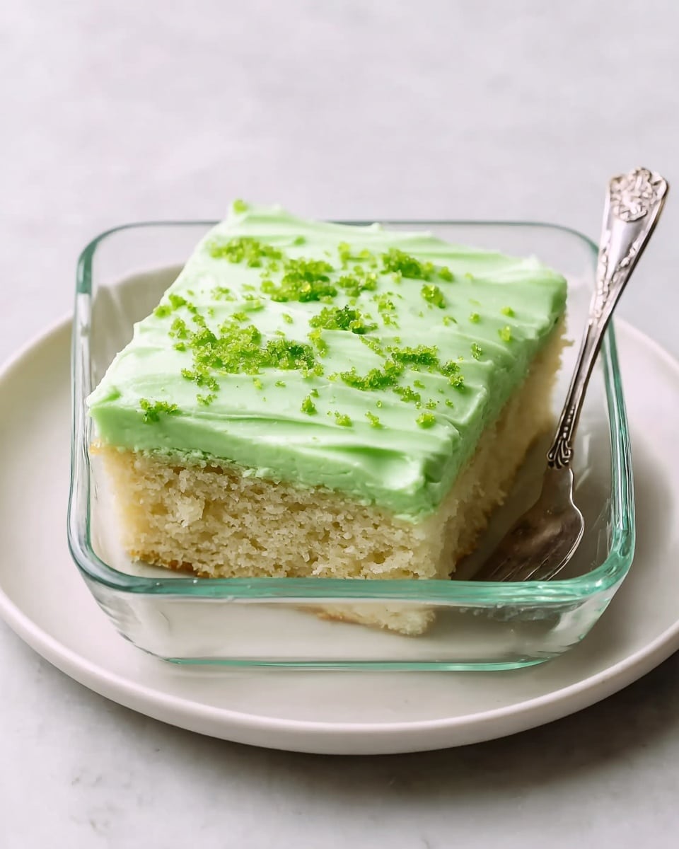 A single square piece of soft, light green cake sits in a clear glass square dish placed on a white plate, all resting on a white marbled textured surface. The cake has one layer of moist, crumbly texture and is topped with a thick, smooth layer of pale green frosting that is spread evenly with gentle waves. Small bright green crumbs are scattered across the top of the frosting, adding a speckled texture. A silver spoon with a detailed handle is placed beside the cake inside the dish. Photo taken with an iphone --ar 4:5 --v 7