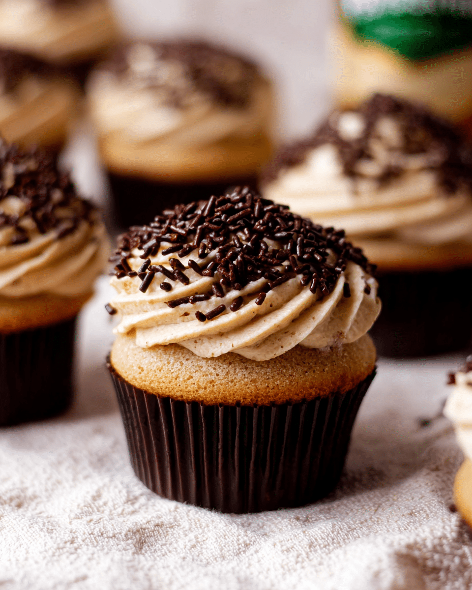 The image shows a close-up of a cupcake with two main layers: the bottom layer is a light brown cake in a dark brown paper wrapper, and the top layer is a thick swirl of creamy, tan-colored frosting covered generously with dark brown chocolate sprinkles. There are multiple cupcakes of the same kind slightly out of focus in the background, all placed on a light textured cloth with a white marbled backdrop. The focus is sharp on the cupcake in the center, emphasizing the texture of the cake and the smooth, creamy frosting dotted with sprinkles. photo taken with an iphone --ar 4:5 --v 7