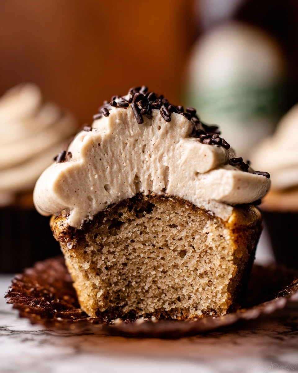 The image shows a close-up of a cupcake cut in half, revealing two main layers: the bottom layer is a moist, light brown cake with a soft, slightly crumbly texture, and the top layer is a thick swirled light beige cream frosting, crowned with scattered small dark chocolate sprinkles. The cupcake sits in a dark brown liner, resting on a white marbled surface, with a blurred background creating a warm and cozy feel. Photo taken with an iphone --ar 4:5 --v 7