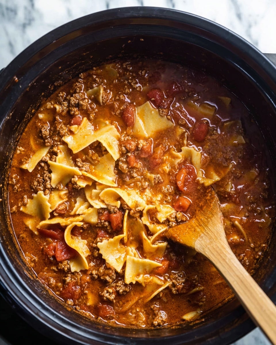 This image shows a black pot filled with a thick stew consisting of a reddish-brown broth, chunky browned ground meat, soft diced red tomatoes, and irregular wide yellow pasta pieces scattered on top. A wooden spatula with some sauce on it rests inside the pot toward the upper right corner. The stew has a glossy, hearty texture with visible bits of seasoning and small pasta shapes mixed throughout. The scene is set on a white marbled surface. Photo taken with an iphone --ar 4:5 --v 7