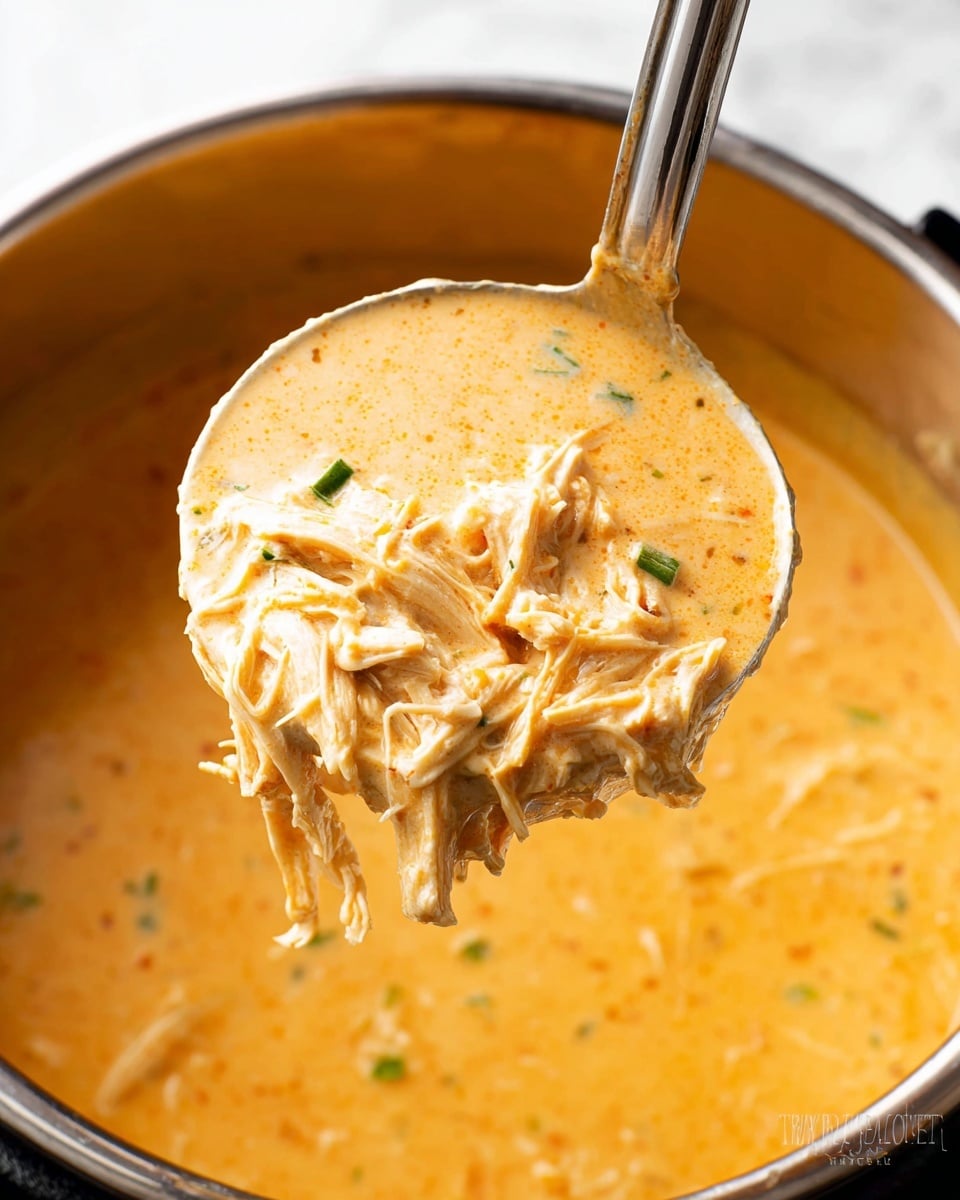 The image shows a close-up of a thick creamy soup with shredded chicken and small green bits, held in a silver ladle above a large pot filled with more of the orange-colored soup. The soup is smooth with visible strands of chicken mixed in, giving it texture. The base is a rich, light orange color with a few small green pieces scattered throughout. The ladle lifts a generous portion showing the dense, creamy texture clearly. The background is a white marbled texture. photo taken with an iphone --ar 4:5 --v 7