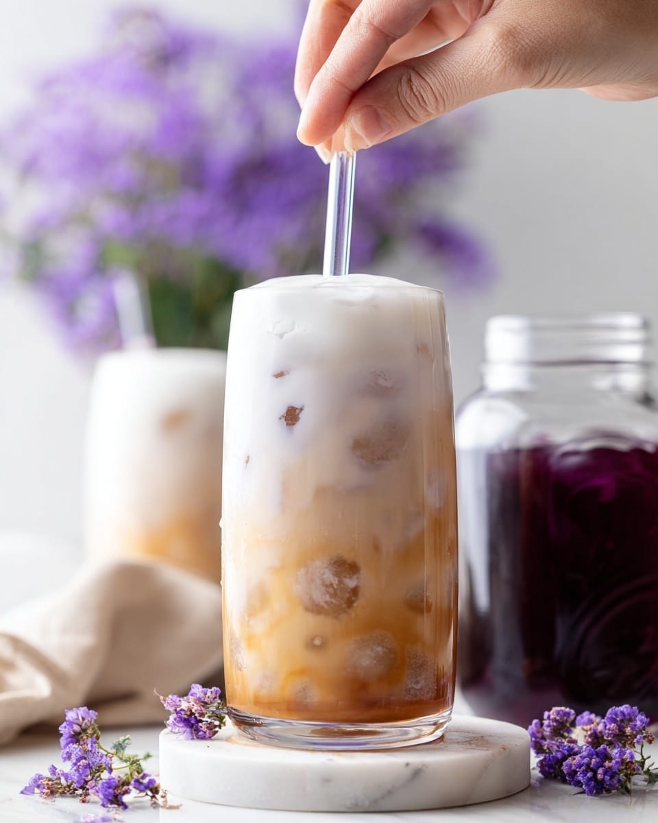 There is a tall clear glass filled with three main layers: the bottom layer is light brown iced milk with scattered translucent ice cubes inside; above it is a thick white foam layer topping the drink; a woman's hand is holding a clear straw that touches the foam peak on top. The glass is placed on a round white coaster over a white marbled texture, with small purple flowers near the base on the right. In the blurred background, another glass with the same drink and a clear jar filled with a deep purple liquid are visible along with a bunch of purple flowers. Photo taken with an iphone --ar 4:5 --v 7