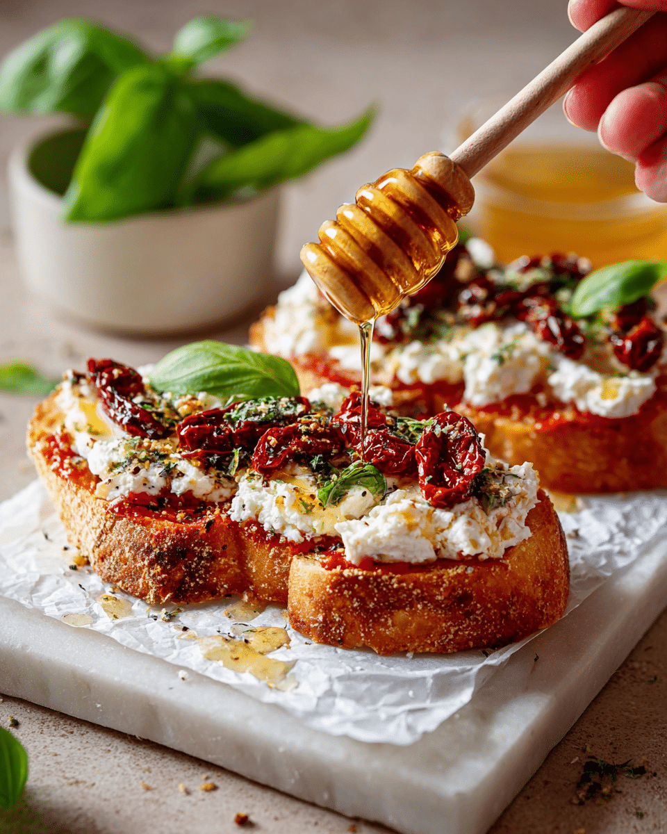 Three pieces of toasted bread lie on brown parchment paper on a silver tray. Each toast has a base layer of red tomato sauce, topped with a thick layer of white, crumbly cheese spread. On top, dark red sun-dried tomatoes are spread unevenly, with bright green fresh basil leaves scattered over all. The toasts have a rustic texture with golden edges. Around the tray, there are fresh basil leaves, slices of plain bread, a small dish of honey with a wooden dipper, and a bowl of dried herbs on a white marbled surface. photo taken with an iphone --ar 4:5 --v 7
