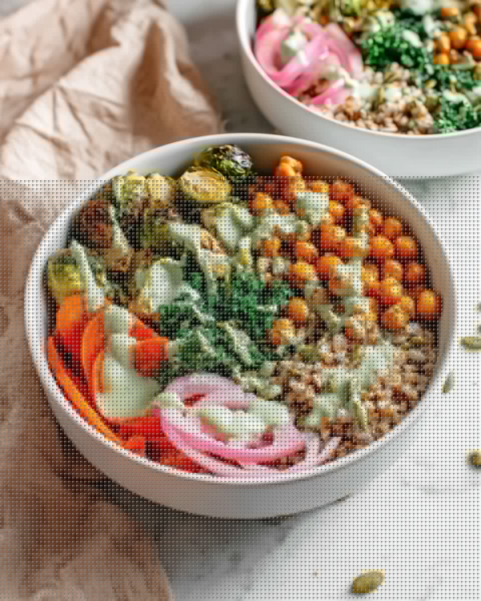 A white bowl filled with layers of healthy food starting with a base of brown grains at the bottom right, topped with cooked orange chickpeas covered in a light green creamy sauce. On the top left, there are roasted green broccoli and brussels sprouts with the same creamy drizzle. In the center, bright green chopped kale is visible, and on the bottom left, cooked sliced orange carrots add a bright contrast. On the bottom right, there are thin, pale pink pickled red onion rings, and pumpkin seeds are scattered on top of the whole bowl. Another similar bowl is partially visible in the background, sitting on a white marbled surface with a soft beige cloth nearby. photo taken with an iphone --ar 4:5 --v 7