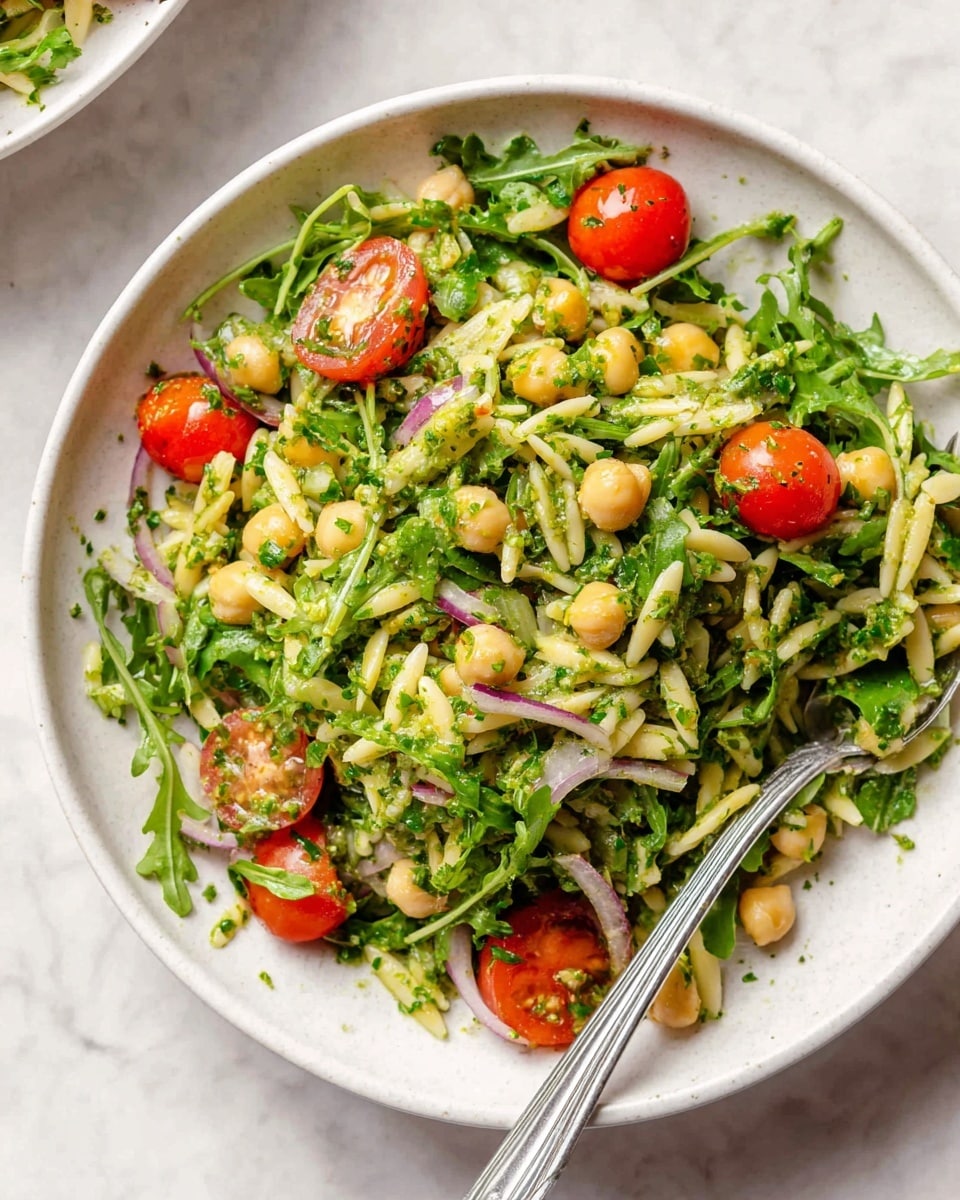 A white bowl filled with a fresh salad on a white marbled textured surface, showing layers of bright green leafy arugula and parsley mixed with beige chickpeas and light yellow orzo pasta, scattered with whole and halved shiny red cherry tomatoes, and thin slices of pale purple red onion. The salad looks lightly coated in a green herb dressing and there is a silver fork resting inside the bowl. Photo taken with an iphone --ar 4:5 --v 7