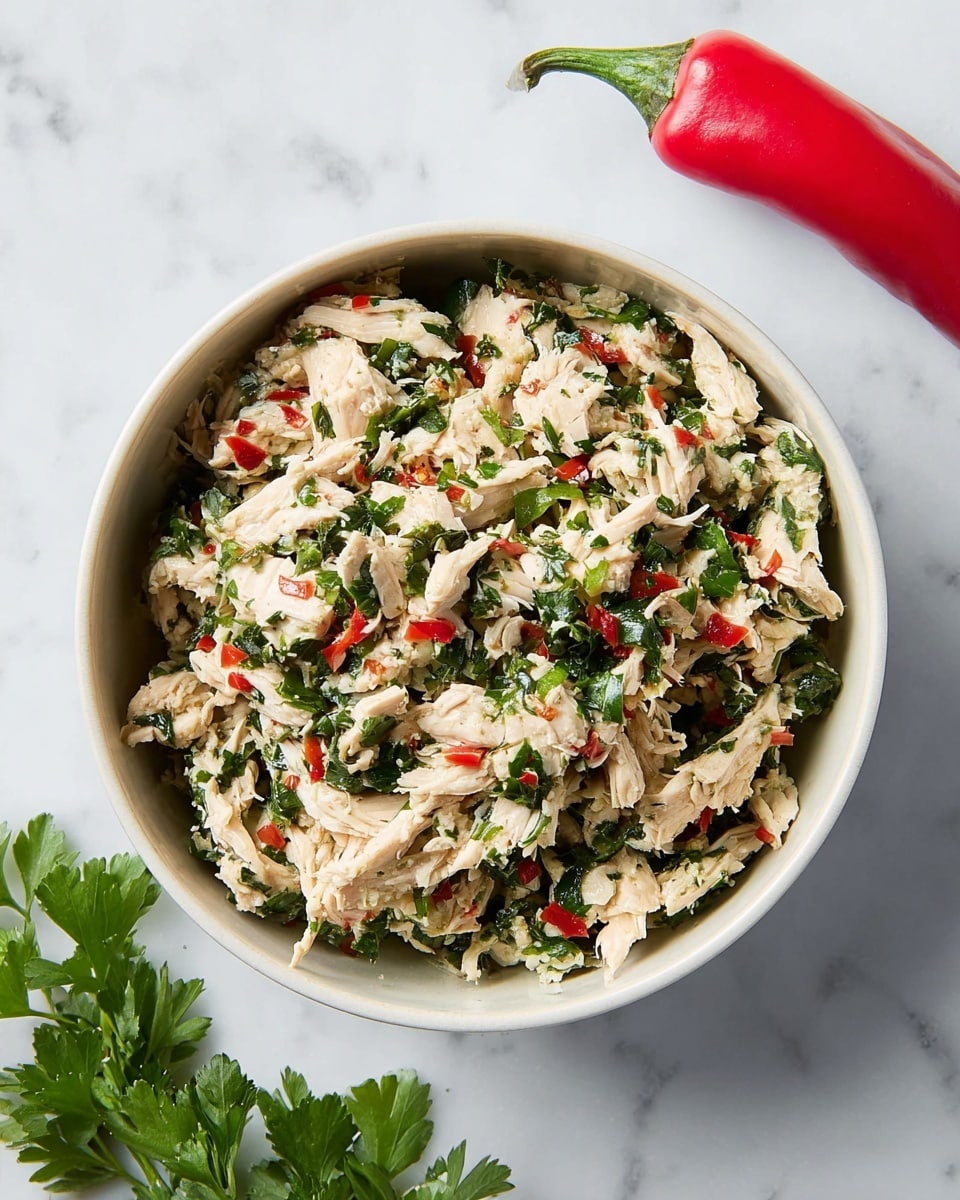 A top-down view of a single-layer white bowl filled with a mixture of white shredded chicken, chopped vibrant green herbs, and small bits of red chili. The chicken pieces look tender with a slightly moist texture, mixed evenly with fresh leafy green herbs and scattered red chili pieces for contrast. The bowl sits on a white marbled surface alongside a whole red chili pepper with a green stem in the top right corner and a sprig of green parsley in the bottom left corner, adding fresh elements around the dish. photo taken with an iphone --ar 4:5 --v 7