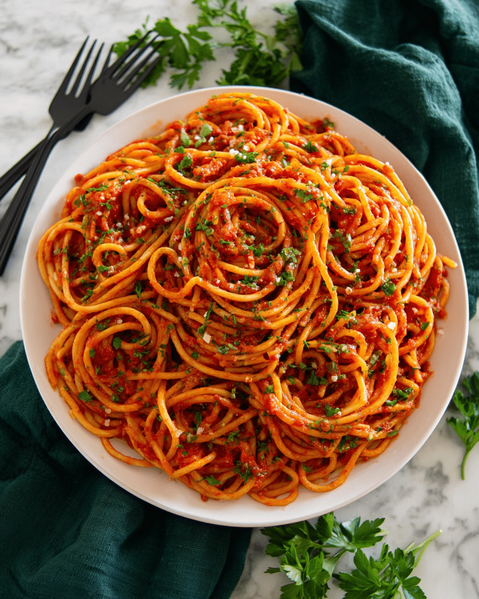 A round white plate filled with a single layer of thick spaghetti noodles covered in rich red tomato sauce mixed with small bits of garlic and herbs; the pasta is arranged in loose swirls and sprinkled with chopped green parsley pieces that add a fresh contrast, placed on a dark green cloth on a white marbled surface with a black fork and spoon in the background, and some parsley leaves scattered nearby. photo taken with an iphone --ar 4:5 --v 7