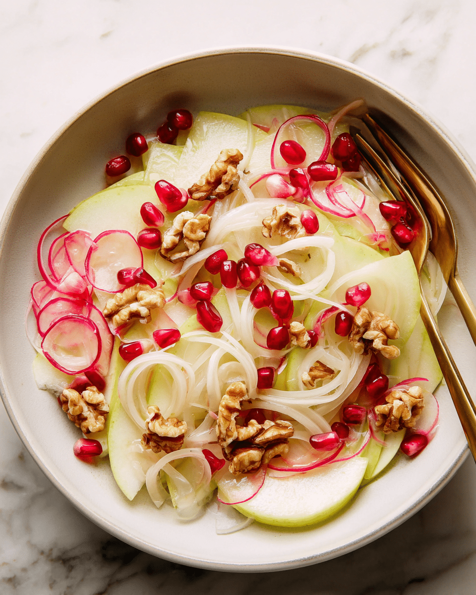 A close-up view of a fresh salad served in a white bowl, with thin slices of green apple and red radish layered at the bottom, topped with pale translucent slices of fennel and rings of light pink pickled onions. Scattered on top are glossy red pomegranate seeds and small clusters of toasted walnuts, adding texture and color contrast. A gold fork is partially visible on the right side of the bowl, resting on the salad. The bowl is placed on a white marbled surface. photo taken with an iphone --ar 4:5 --v 7