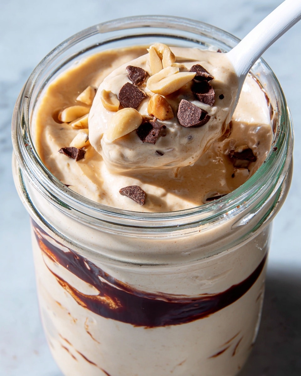 The image shows a close-up of a glass jar filled with a creamy, light beige mixture that has a smooth texture. Inside the jar near the top, there is a visible layer of dark brown sauce swirled on the inside of the glass. On top of the creamy mixture, there are chunks of light tan nuts and dark brown small pieces, likely chocolate bits. A white spoon dips into the jar, lifting a spoonful of the creamy mixture with nuts and chocolate pieces on it. The jar sits on a white marbled surface. Photo taken with an iphone --ar 4:5 --v 7