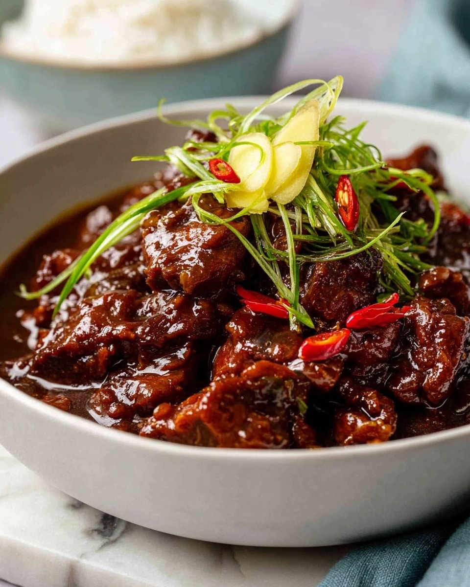 A white bowl filled with a rich, dark brown meat stew that has a glossy, thick sauce coating tender chunks of meat. The stew is topped with thin, curly slices of bright green scallions and a few small, vibrant red chili pieces, along with pale, thin strips of ginger placed on top. The bowl sits on a white marbled surface, with a soft blue cloth nearby and a blurred white bowl of rice in the background. The overall scene is close-up, highlighting the texture and shine of the stew. photo taken with an iphone --ar 4:5 --v 7
