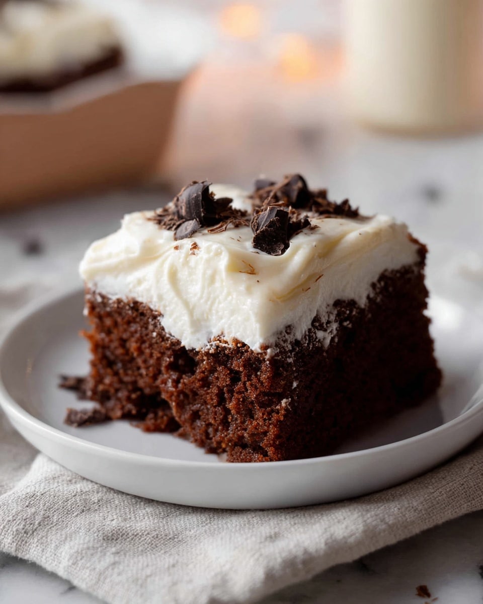 A single piece of chocolate cake sits on a white plate, consisting of two visible layers: a dark brown, moist chocolate base with a soft and airy texture, topped with a thick, creamy white frosting that has a smooth texture and slightly uneven spread. Small dark chocolate curls rest on top of the frosting, adding a decorative touch. The plate is placed on a light beige cloth, and the background shows a blurred white marbled surface with warm, soft lighting. Photo taken with an iphone --ar 4:5 --v 7