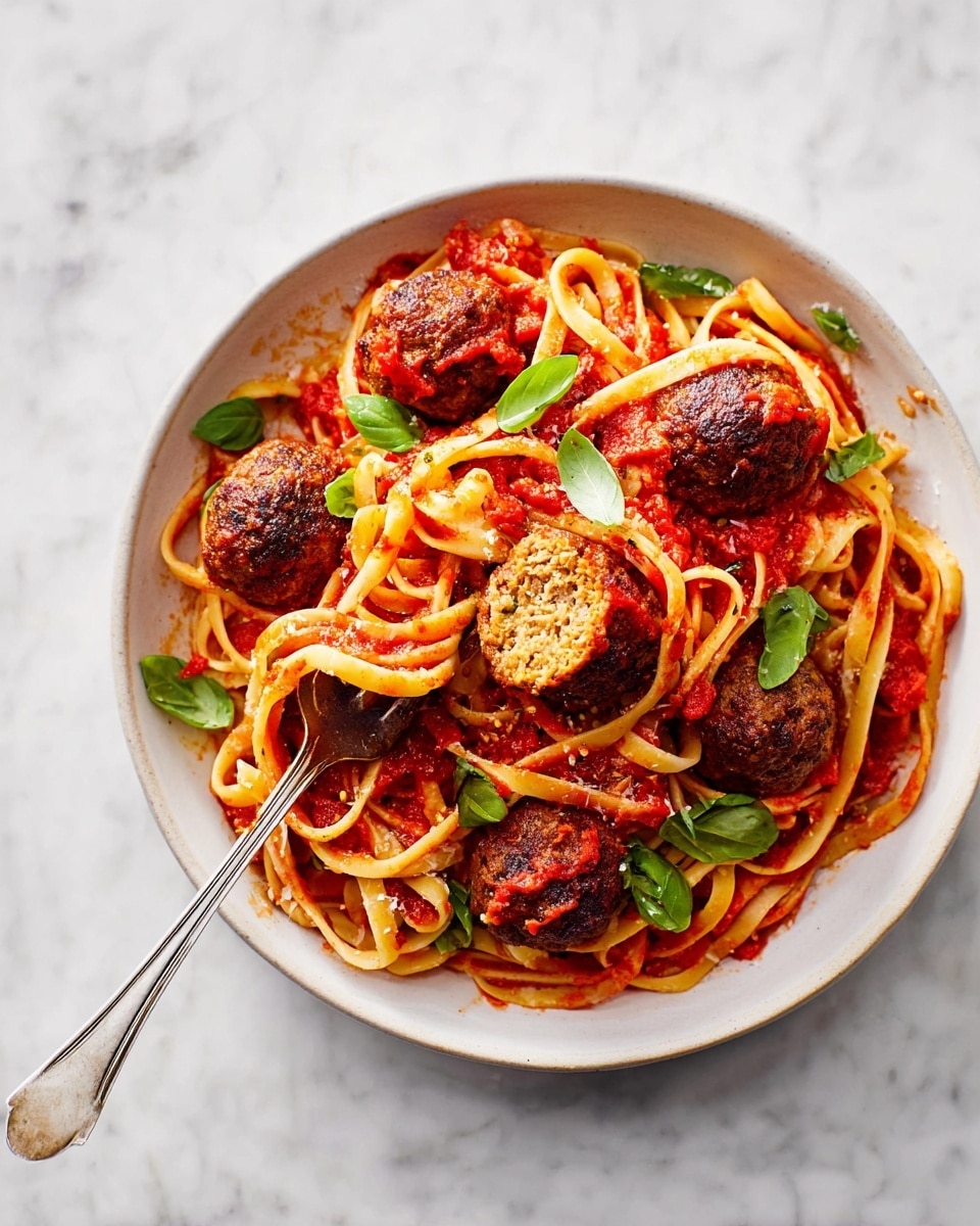 A white bowl holds a serving of linguine pasta mixed with red tomato sauce, topped with six round, brown meatballs, one of which is cut open showing a crumbly, textured inside. Bright green fresh basil leaves are scattered over the pasta and meatballs. A silver fork is twirling some pasta on the right side of the bowl. The dish is set on a white marbled surface. photo taken with an iphone --ar 4:5 --v 7