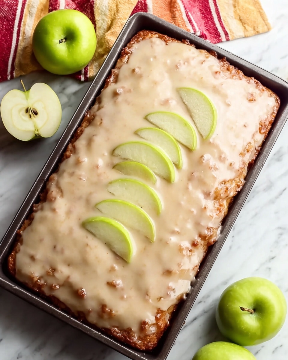 A rectangular cake with one thick layer covered in a glossy, light beige glaze that has a slightly uneven texture with some small lumps. On top, near the middle, are four thin slices of green apple slightly overlapping each other. The cake is in a dark baking tray, set on a white marbled surface, with some whole and halved green apples in the background, along with a folded striped cloth in red, cream, and yellow colors. Photo taken with an iphone --ar 4:5 --v 7