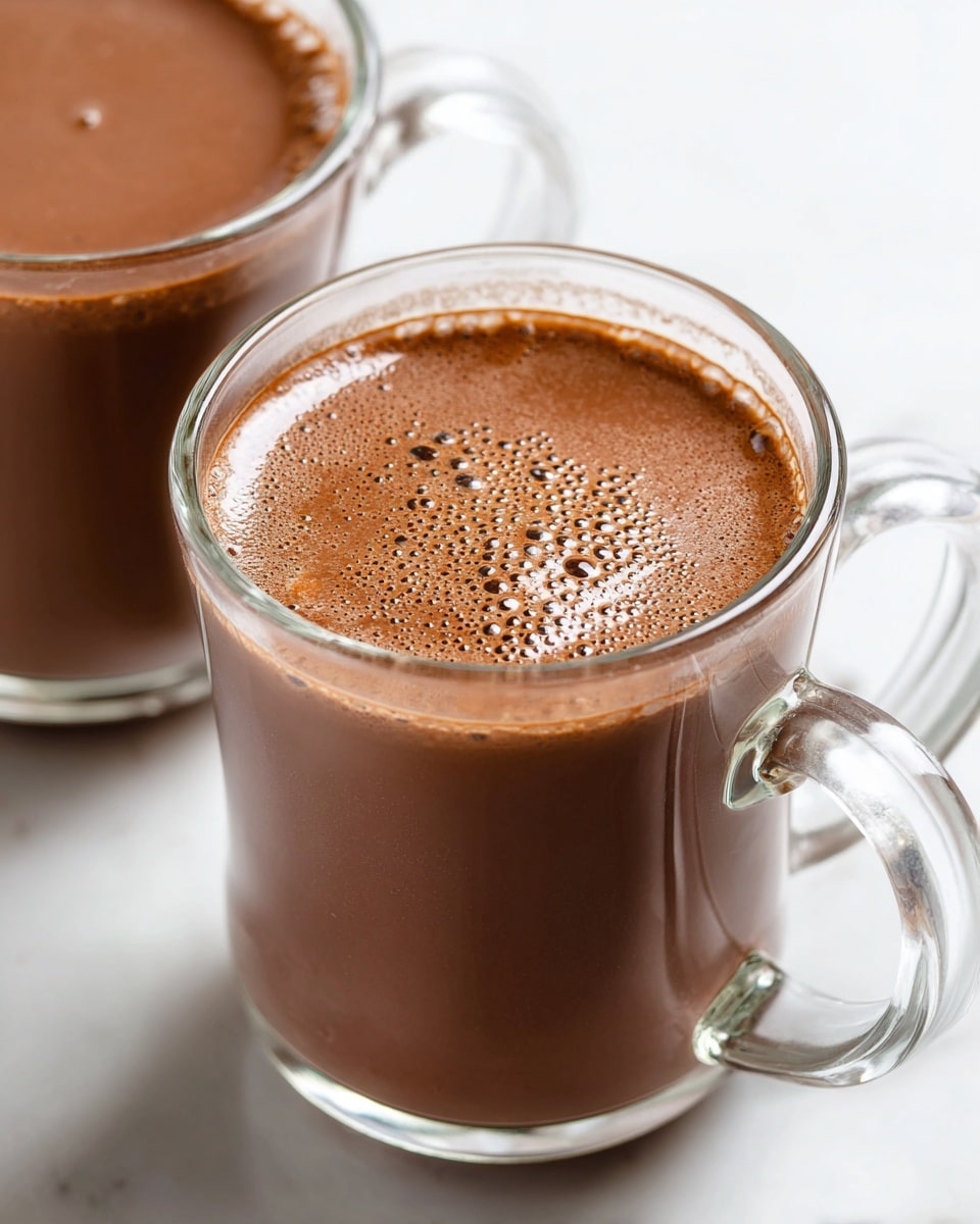 There are two clear glass mugs filled with smooth, rich chocolate drink, showing a dark brown color with small bubbles on the surface. The closest mug is centered and fills most of the frame, while the second mug is blurred in the background on the right side. Both mugs have clear handles and are set on a white marbled surface that brightens the scene. The thick texture and milky shine of the chocolate drink are visible in both mugs. photo taken with an iphone --ar 4:5 --v 7