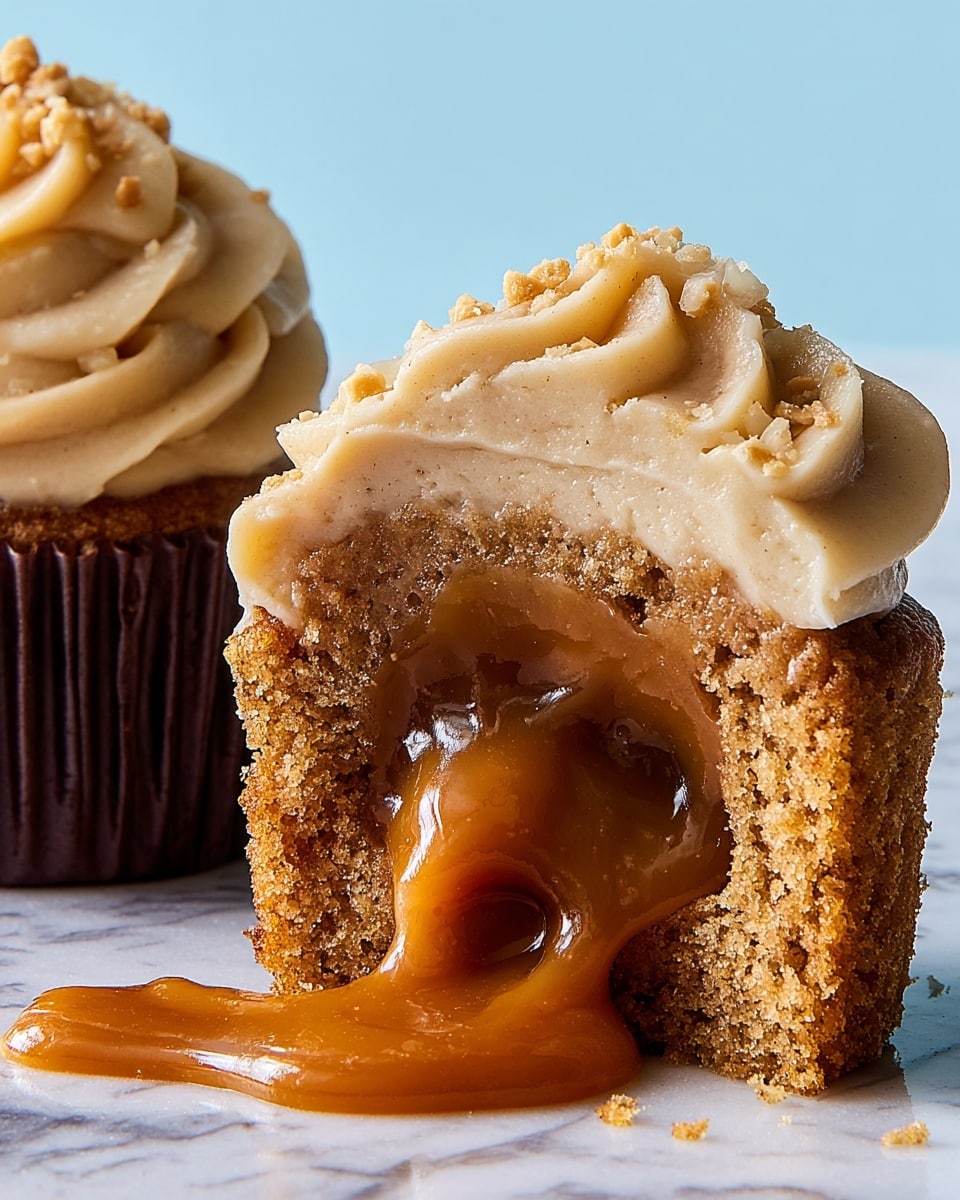 A close-up view of a cupcake cut in half showing three clear layers: the bottom layer is a crumbly, moist, light brown cake; the middle layer is a thick, glossy, flowing caramel sauce spilling onto a white marbled surface; the top layer is a thick, creamy, light tan frosting with a smooth but slightly textured swirl shape, sprinkled with small chopped nuts. Next to it is a whole cupcake in a dark brown paper cup with matching frosting seen on the left edge. The background is a soft, light blue color. Photo taken with an iphone --ar 4:5 --v 7