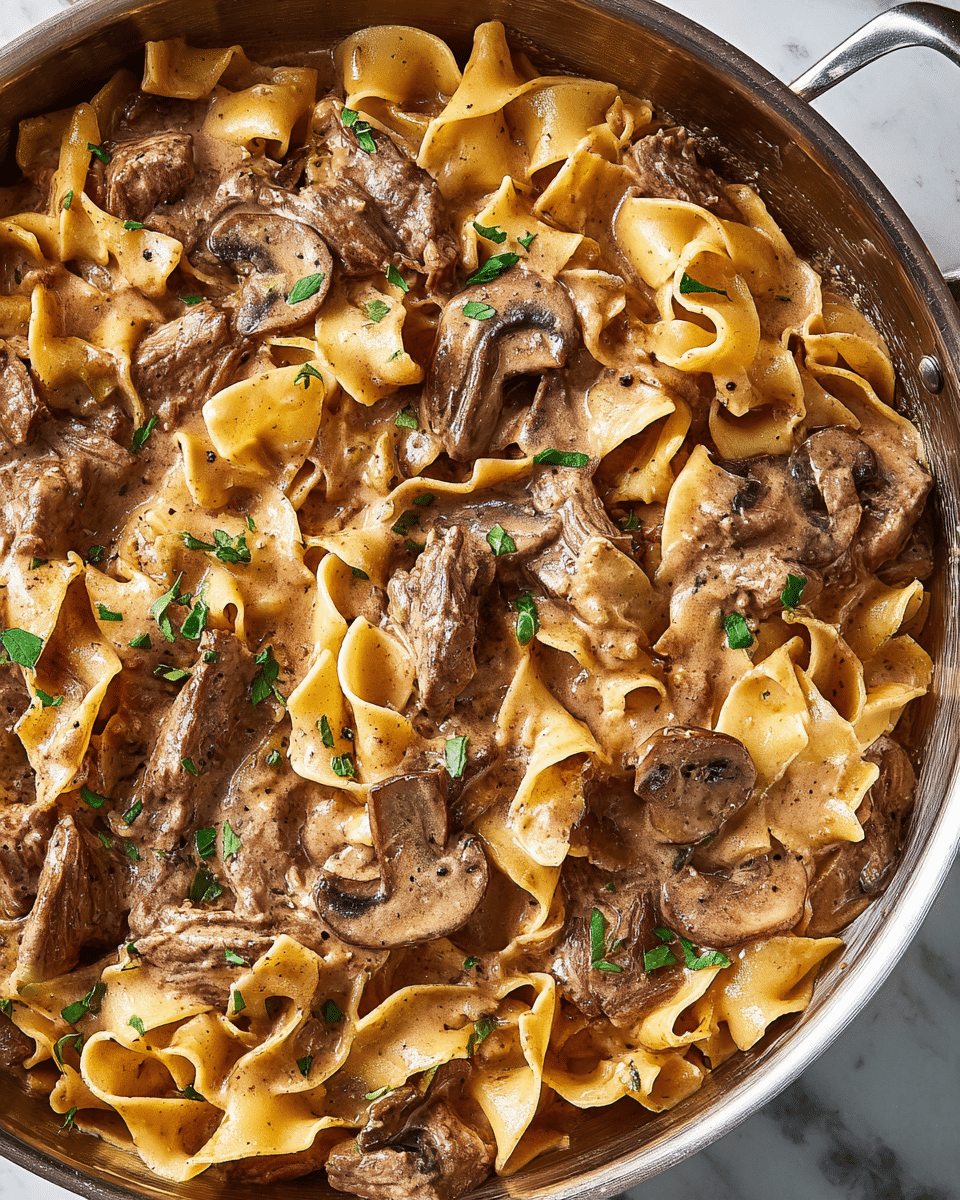 The dish shows wide, flat pasta noodles mixed with tender pieces of beef, all coated in a creamy light brown sauce with visible specks of pepper. There are slices of sautéed mushrooms adding a dark brown element. The pasta and beef layers alternate, with the sauce fully covering everything for a glossy look. Small green herb pieces are sprinkled on top, adding fresh color contrasts. The food is inside a shiny metal pan, and the background is a white marbled texture. photo taken with an iphone --ar 4:5 --v 7