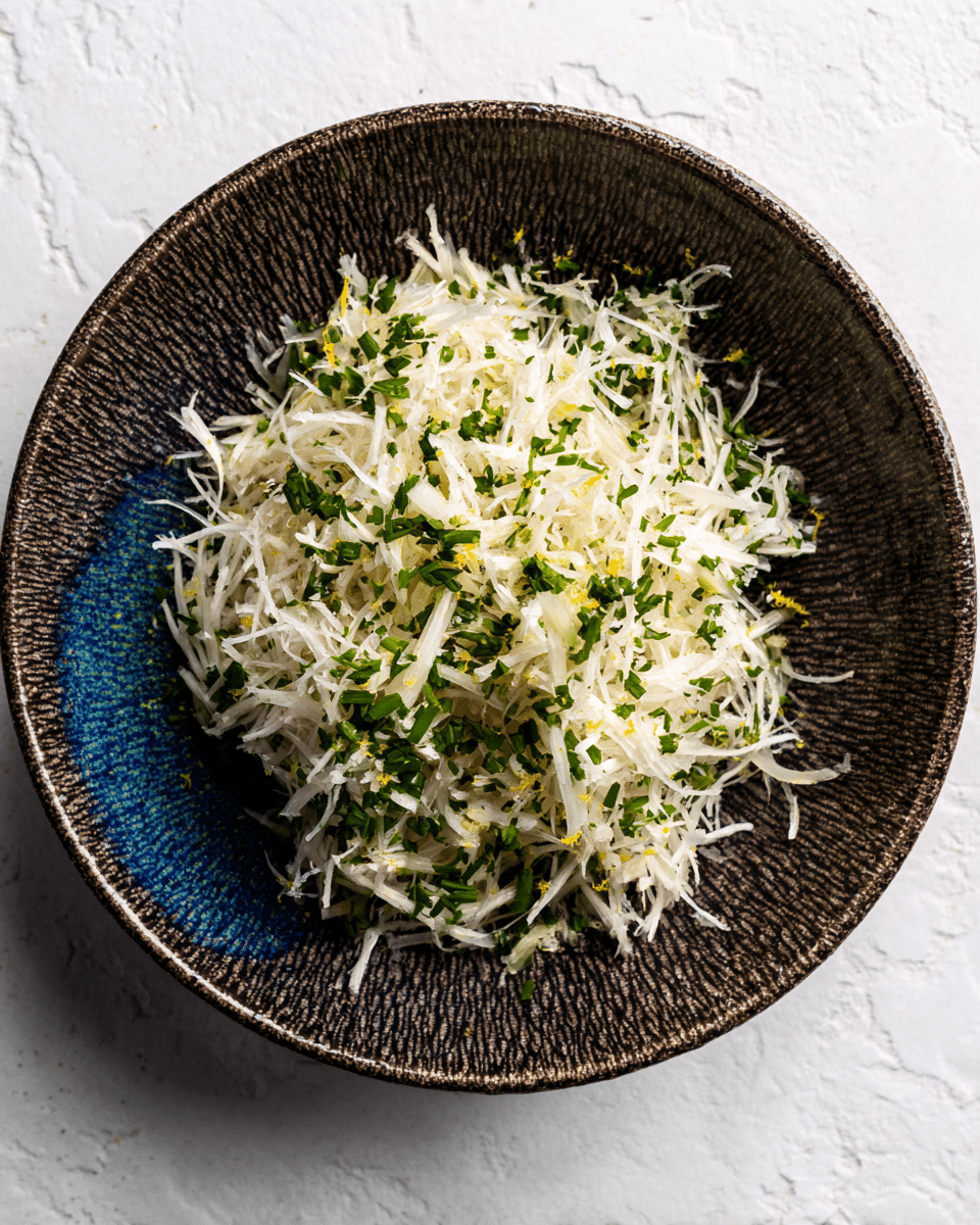 A small mound of thin, white shredded vegetable strands, mixed with finely chopped green herbs and small bits of pale yellow zest, is placed in the center of a textured dark brown bowl with a hint of blue near the rim, sitting on a white marbled surface. The pile looks fresh and light with the green and yellow pieces scattered evenly through the white strands, giving a fresh and natural look. photo taken with an iphone --ar 4:5 --v 7