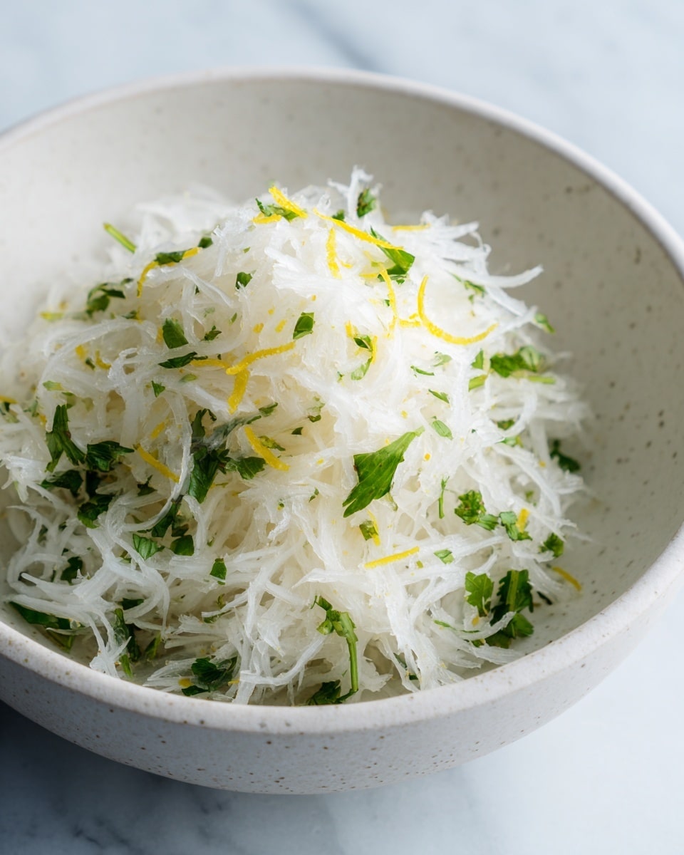 The image shows a close-up of a small mound of thinly shredded white radish mixed with finely chopped green herbs, likely cilantro or parsley, and a few thin yellow zest strips spread lightly on top. This mixture sits in a deep white bowl with a slightly rough texture inside. The radish strands are fresh and crisp with a slightly translucent look, creating a light and refreshing appearance. The bowl is placed on a white marbled surface, providing a clean and elegant background. photo taken with an iphone --ar 4:5 --v 7