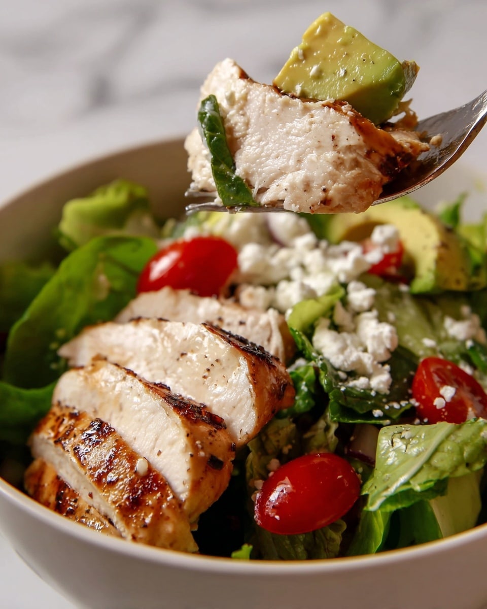 A close-up view of a white bowl filled with a salad made of three layers: at the bottom, thick slices of grilled chicken breast with golden brown grill marks and a moist texture; in the middle, a mix of fresh green lettuce leaves with bright red cherry tomato halves scattered throughout; and on the top, a fork holding a bite showing a piece of grilled chicken, a chunk of creamy green avocado sprinkled with white feta cheese crumbles, a cherry tomato half, and some leafy spinach. The background is a white marbled texture. Photo taken with an iphone --ar 4:5 --v 7