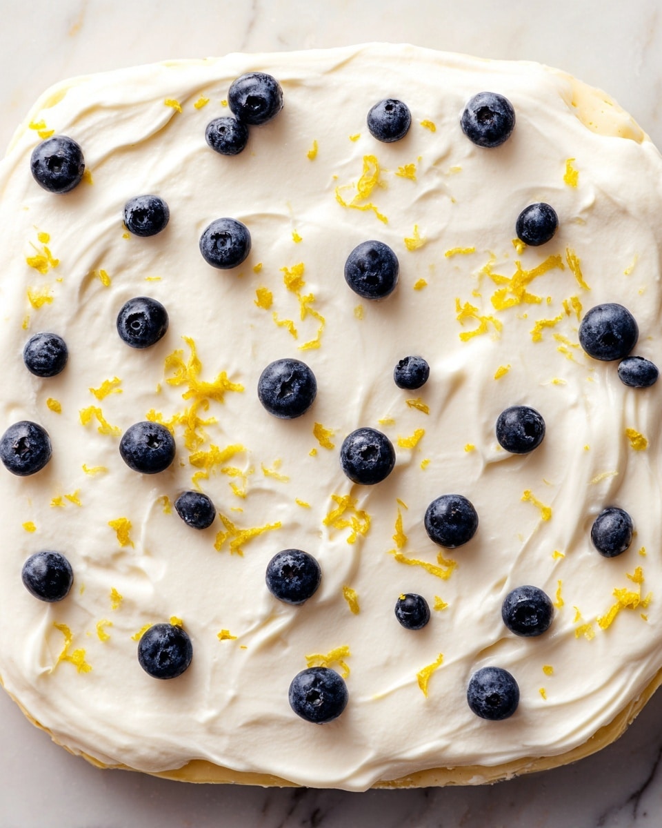 A single flat layer dessert with a pale yellow base topped with a thick, smooth white cream spread evenly over it. Scattered across the cream are small, round, dark blue blueberries and tiny bright yellow lemon zest pieces that add contrast. The edges of the dessert are slightly visible, showing the pale base under the cream. The background is a white marbled surface. photo taken with an iphone --ar 4:5 --v 7