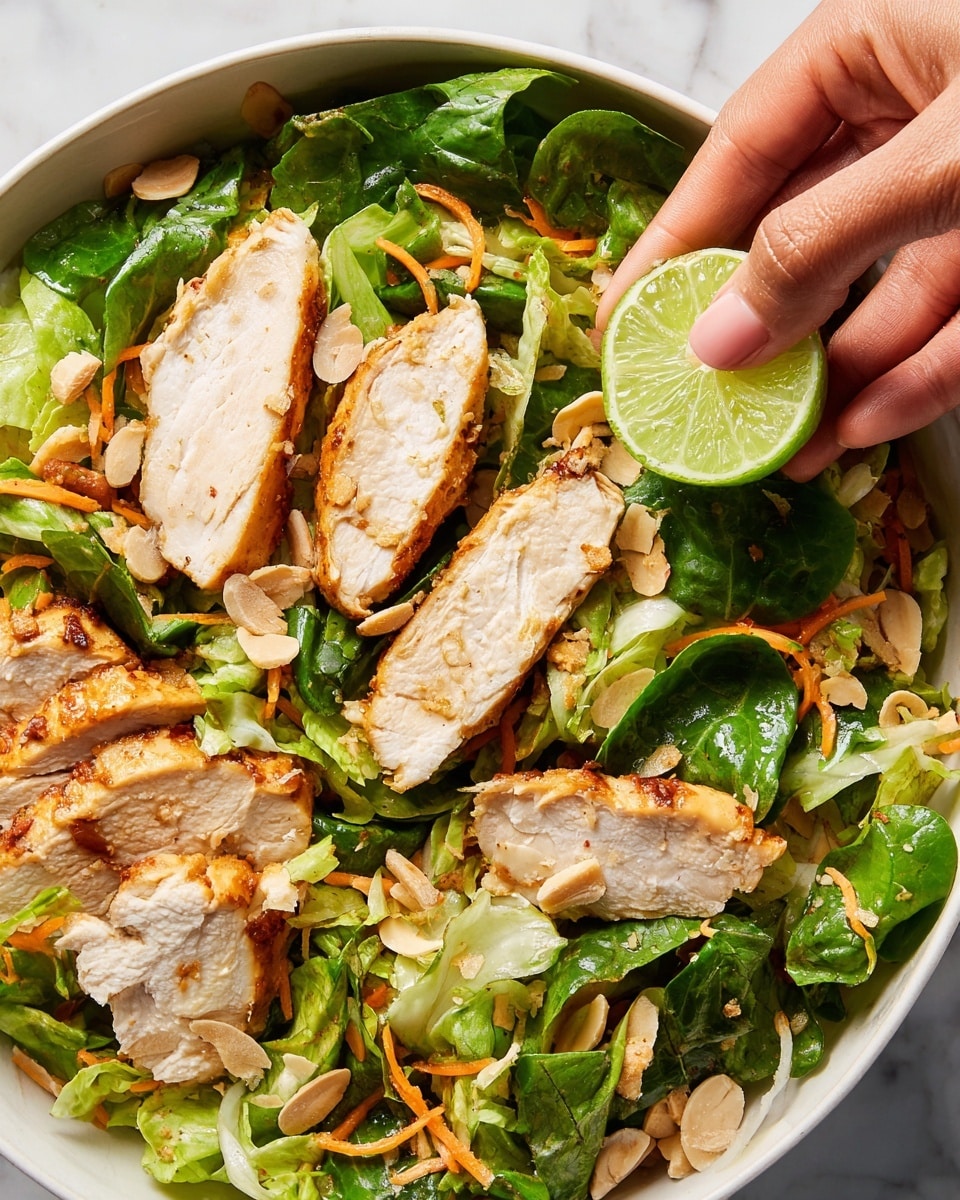 A close-up view of a white bowl filled with a fresh salad showing several thick slices of golden-brown cooked chicken placed on a bed of bright green lettuce and shredded orange carrots. Scattered on top are small pieces of light beige nuts or coconut flakes adding texture. A woman's hand is holding a small wedge of green lime over the salad, ready to squeeze it. The bowl sits on a white marbled background. Photo taken with an iphone --ar 4:5 --v 7