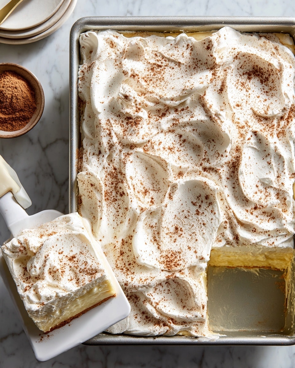 A rectangular metal baking pan filled with a dessert that has two visible layers: a bottom yellowish creamy layer and a thick top layer of whipped white cream, swirled with soft peaks and sprinkled evenly with brown cinnamon powder. A square piece is cut out and lifted on a white spatula, showing the same two layers clearly, with the top whipped cream layer thick and textured. The pan is on a white marbled surface with a small bowl of cinnamon powder and white plates nearby. photo taken with an iphone --ar 4:5 --v 7