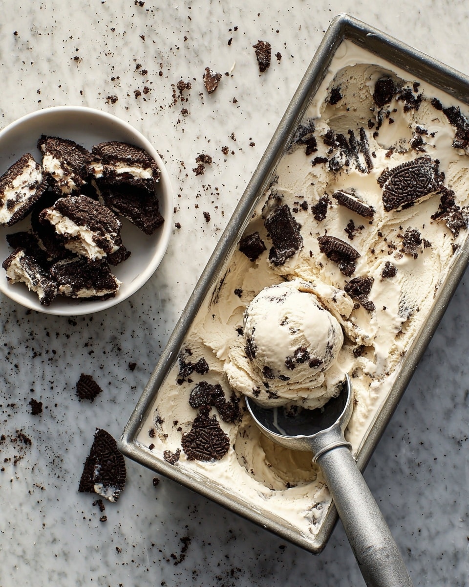 A rectangular metal tray filled with creamy white ice cream mixed with broken pieces of dark chocolate cookies scattered evenly throughout the smooth texture forms the main layer. On the right side of the tray, a scoop of this ice cream is lifted, showing a chunky mix of cookie pieces held together by the soft ice cream. To the upper left next to the tray, a white bowl holds more broken cookie pieces with a rough dark brown texture and white filling visible in some parts. The whole setting is on a white marbled surface with bits of cookie crumbs scattered around. photo taken with an iphone --ar 4:5 --v 7