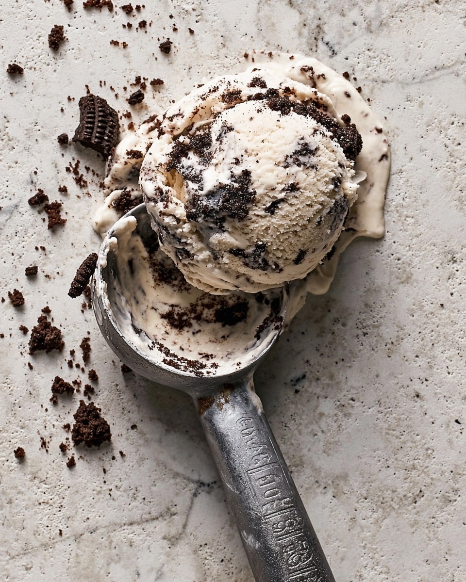 A single scoop of cookies and cream ice cream, creamy white with dark chocolate cookie chunks mixed throughout, sits slightly melting on a white marbled textured surface. The ice cream has a soft, smooth texture with bits of cookie giving a rough, crumbly look on top and around the scoop. Scattered cookie crumbs in dark brown are visible around the scoop. Next to it lies a metal ice cream scoop with some melted ice cream and cookie residue inside and on its edges, all resting on the same white marbled texture. photo taken with an iphone --ar 4:5 --v 7