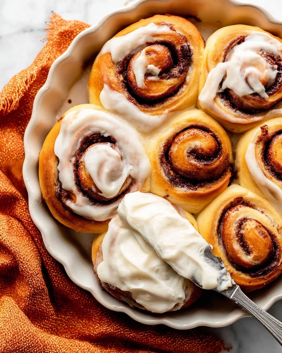 The image shows a close-up of seven cinnamon rolls in a white ceramic round dish with scalloped edges, placed on a white marbled surface with a textured orange cloth nearby. Each cinnamon roll has a golden brown dough base with a dark brown cinnamon sugar swirl inside, creating a spiral pattern. Four of the rolls have thick, creamy off-white icing generously spread over their tops, some showing specks of vanilla. A metal spreading knife with icing on it rests on one cinnamon roll at the bottom right, as if in the middle of spreading. The texture of the dough looks soft and fluffy with a slight shine, while the cinnamon filling appears dense and moist. Photo taken with an iphone --ar 4:5 --v 7