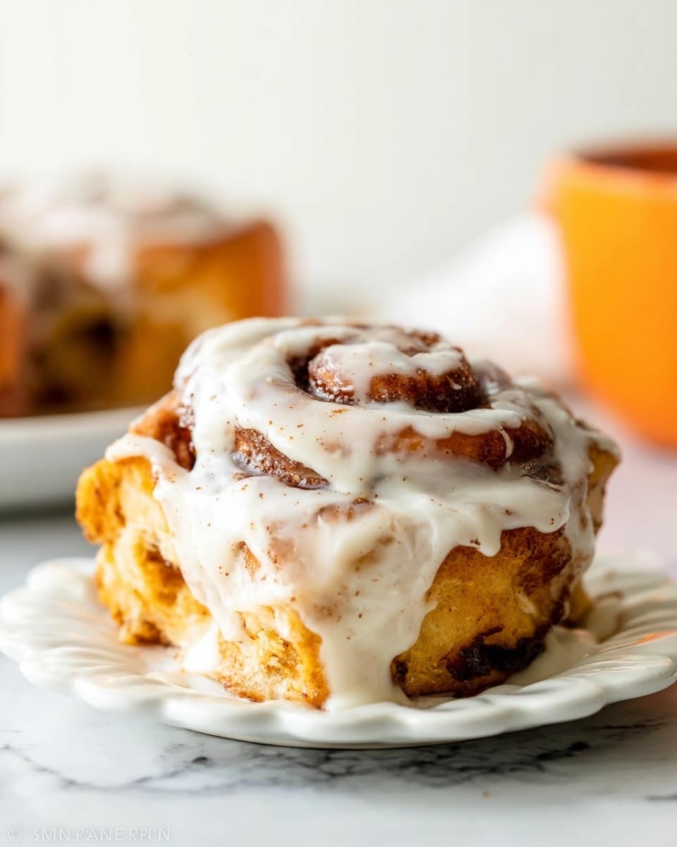 A close-up of a single cinnamon roll on a white plate with a scalloped edge, showing three visible layers of soft, golden-brown dough rolled with dark cinnamon sugar. The roll is generously covered with a thick, creamy, off-white icing that drips down the sides, rich in texture with visible specks of cinnamon. The background is a white marbled texture with a blurred second cinnamon roll and a soft orange shape to the right. The lighting is bright and natural, highlighting the glossy icing and moist dough. photo taken with an iphone --ar 4:5 --v 7