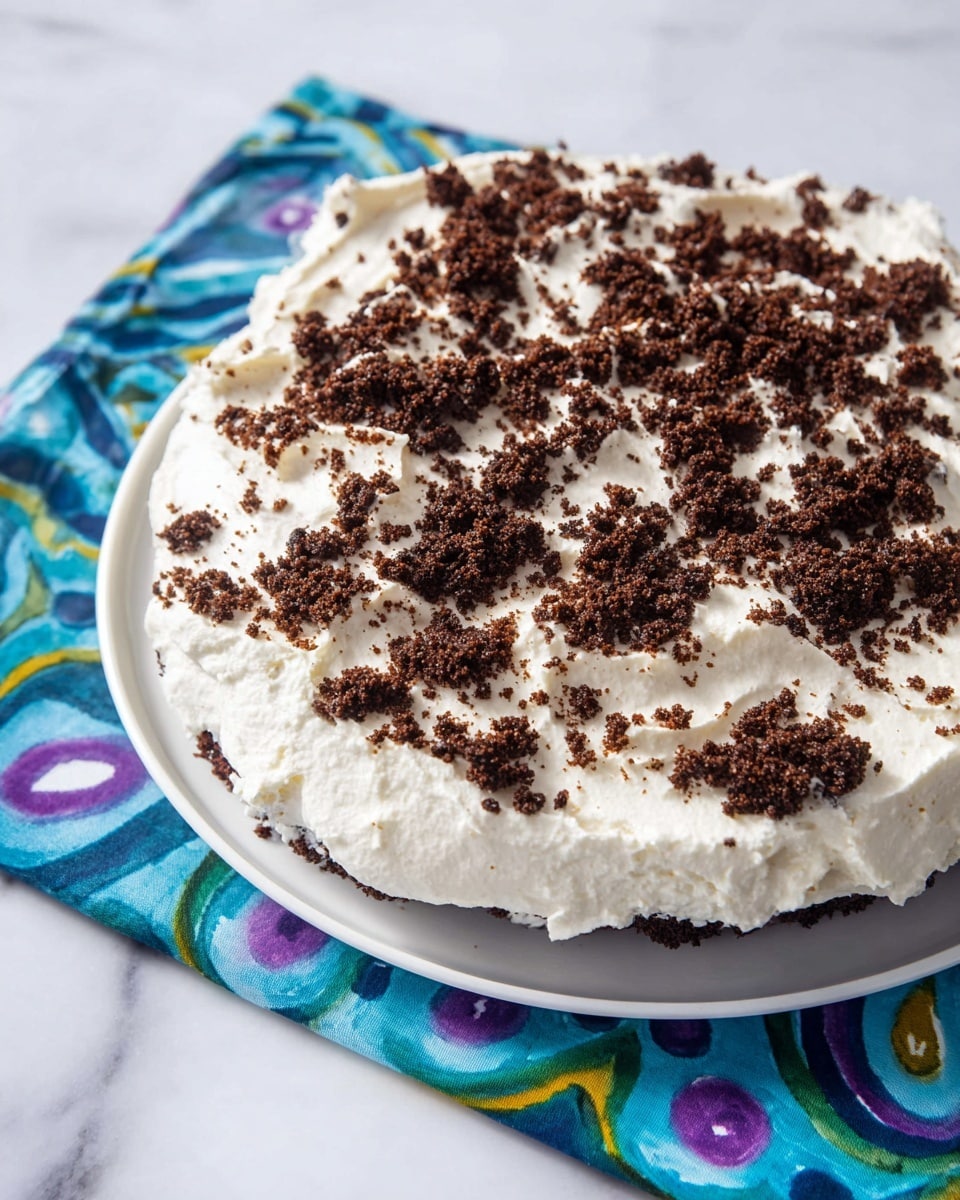 The dish is a cake with one visible layer of white whipped cream spread evenly on top, creating a soft, smooth texture. Scattered across the whipped cream are small, rough chunks of dark brown chocolate cake crumbs, adding a contrast in color and texture. The cake sits on a white plate with a colorful blue, green, and purple patterned cloth underneath it, and the surface beneath is a white marbled texture. photo taken with an iphone --ar 4:5 --v 7