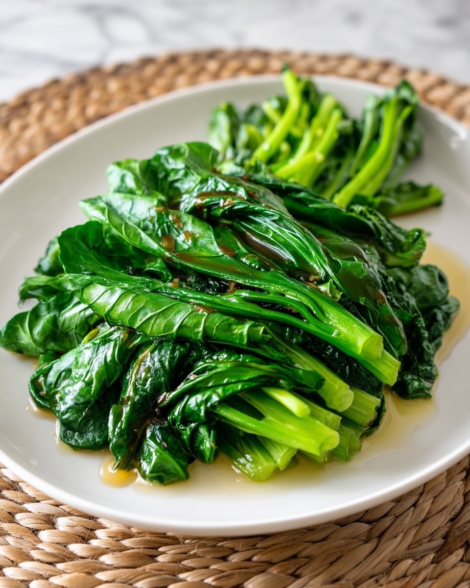 A white plate holds a simple dish of cooked leafy greens, arranged in one main layer that covers most of the plate. The leaves are deep green, slightly shiny from a light sauce drizzled unevenly on top, giving a moist look. The greens appear soft and tender with visible veins and slight curling at the edges. Behind the main leaves, there is a small grouping of bright green stems that add a fresh contrast. The whole plate is placed on a textured, woven mat, but the background is changed to a white marbled texture. photo taken with an iphone --ar 4:5 --v 7