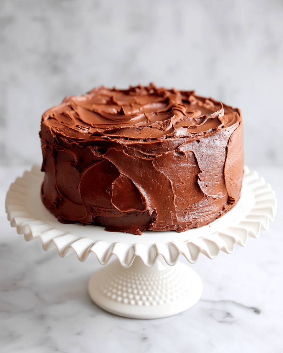 A round chocolate cake with thick, smooth, and creamy chocolate frosting covering the entire surface. The frosting is spread unevenly with visible swirls and peaks, giving it a rustic look. The cake sits on a white cake stand with a textured base, and the background is a white marbled texture. Photo taken with an iphone --ar 4:5 --v 7