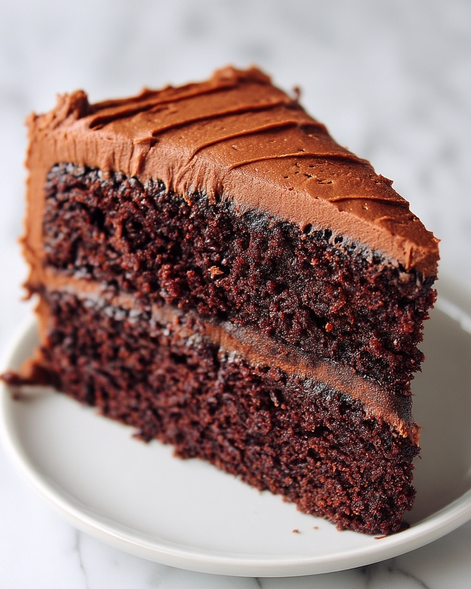 A close-up slice of two-layer chocolate cake sits on a white plate against a white marbled background. The bottom and top layers are moist, dark chocolate cake with a soft crumb texture. Between the two layers and covering the top is smooth, rich, medium brown chocolate frosting, spread thickly with lightly textured strokes visible. The edges of the cake slice show the dense chocolate frosting and moist interior clearly. Photo taken with an iphone --ar 4:5 --v 7