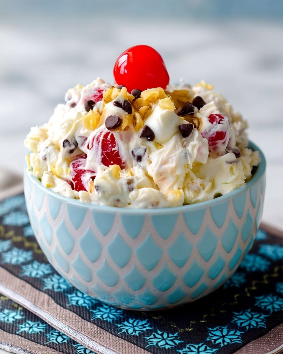 A bowl filled with a creamy white mixture, dotted with bright red cherries and small dark chocolate chips evenly spread throughout. The mixture appears fluffy and thick, with small pieces of light yellow nuts scattered on top, adding texture. A single shiny red cherry sits atop the mound of the mixture as a final garnish. The bowl is white with a light blue fish scale-like pattern and is placed on a layered dark blue and beige patterned cloth, all set against a white marbled background. Photo taken with an iphone --ar 4:5 --v 7