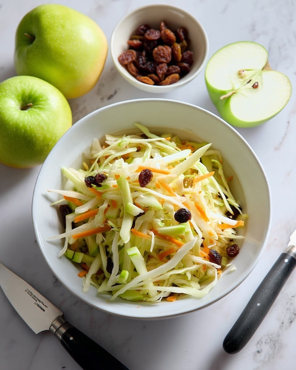 A white bowl holds a fresh salad made of thinly shredded pale green cabbage, light orange carrot strips, and small pieces of green apple with a smoother texture, all mixed with dark brown raisins scattered through the layers. Around the bowl on a white marbled surface, there are two whole green apples, one cut in half revealing its pale green inside, and a white bowl with more raisins. A knife with a black handle lies next to the cut apples. The overall scene is bright with natural light. photo taken with an iphone --ar 4:5 --v 7