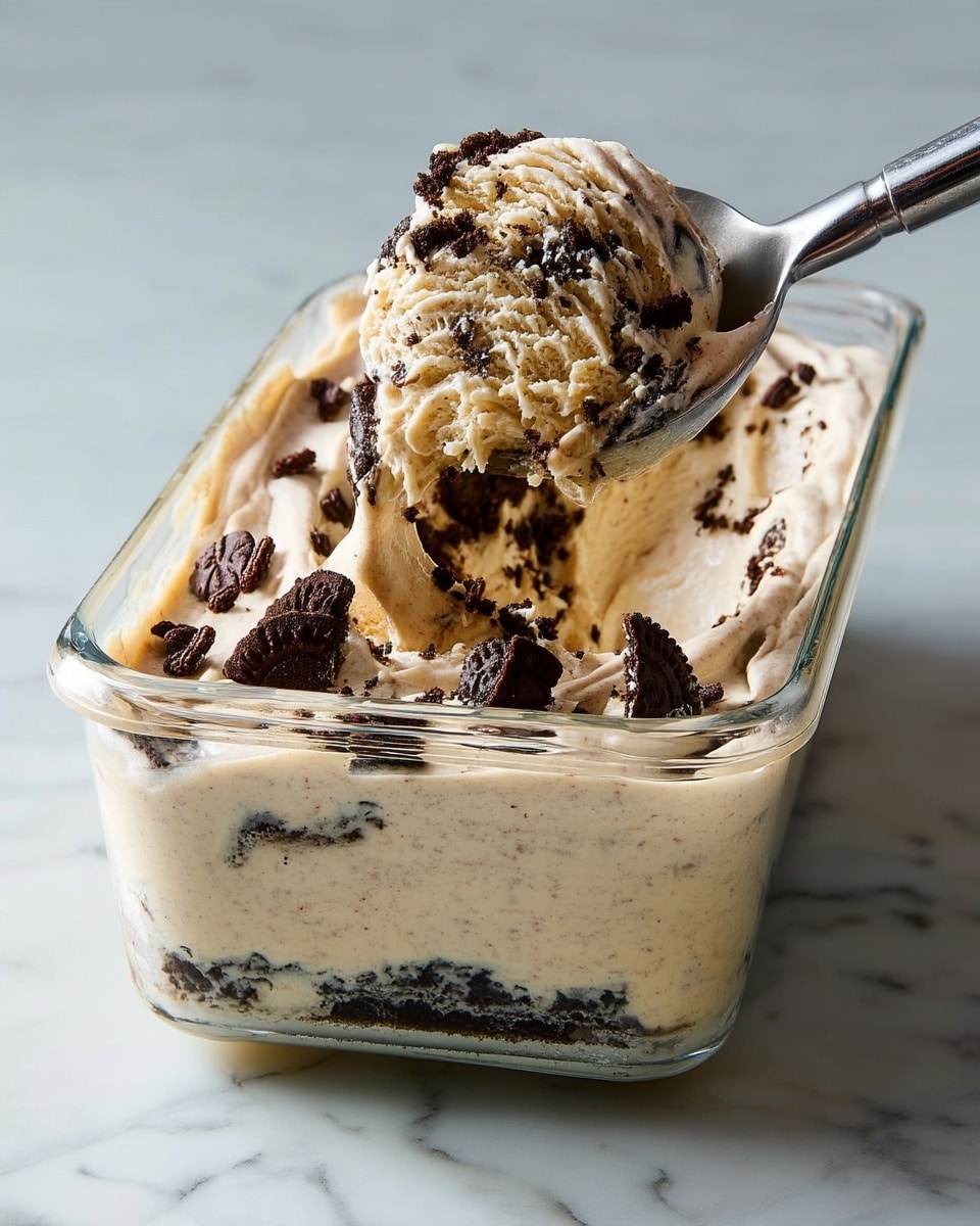 A close-up view of creamy ice cream mixed with dark brown crushed cookie pieces in a clear glass container, showing thick swirls of light beige ice cream with plenty of dark cookie chunks embedded throughout. A metal spoon lifts a scoop filled with the same beige ice cream and broken dark cookie pieces. The container sits on a white marbled texture background. Photo taken with an iphone --ar 4:5 --v 7