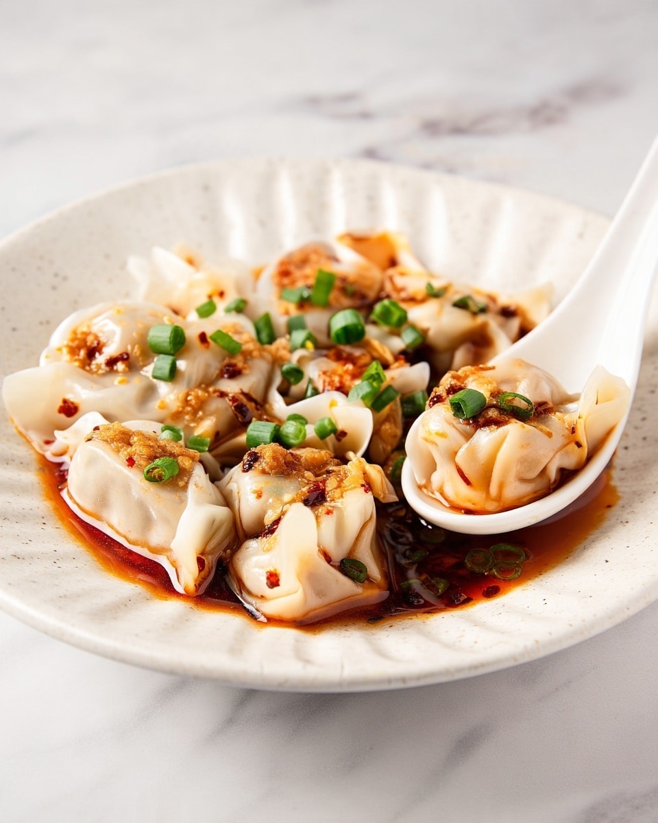 A white plate holds a single layer of round dumplings with thin, translucent wrappers that are slightly folded at the edges, creating a soft, flowing texture. The dumplings are covered with a dark reddish-brown chili oil sauce that pools lightly on the plate. On top, small pieces of golden-brown fried garlic and bright green chopped scallions are sprinkled evenly, adding crispness and color contrast. A white ceramic spoon is nestled on the side of the plate, gently lifting one dumpling with visible folds. The setup is on a white marbled texture surface. photo taken with an iphone --ar 4:5 --v 7