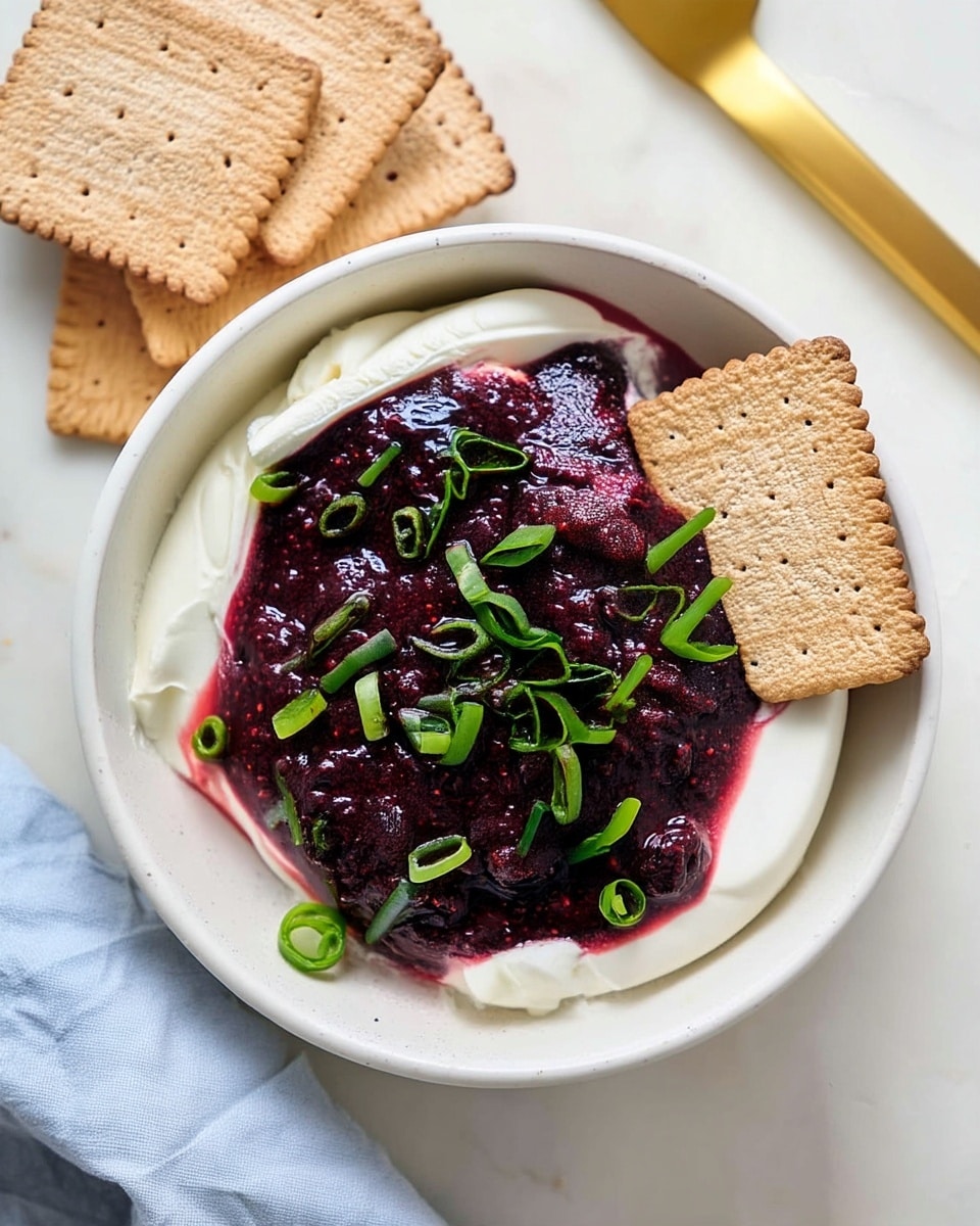 The image shows a white bowl with two main layers: a thick, smooth, white cream layer at the bottom and a thick deep purple berry mixture layer on top. The berry layer has a slightly textured surface with small seeds visible. Green chopped scallions are scattered evenly over the purple layer. A square beige cracker leans on the right edge of the bowl, partially resting on the cream. Three more crackers are stacked off to the top left corner of the bowl, all on a white marbled surface. Near the bowl’s top right is a shiny gold spreader knife. A light blue cloth is partially visible at the bottom left corner. Photo taken with an iphone --ar 4:5 --v 7