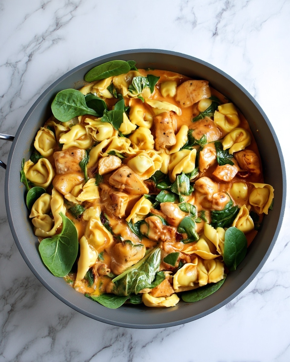 A gray pan is filled with a dish made of three main layers: large pieces of light brown cooked chicken scattered evenly, yellow tortellini pasta with a folded texture filling most of the pan, and fresh green spinach leaves placed on top and mixed throughout; all these are covered in a smooth, creamy orange sauce that fills the spaces between the ingredients. The pan is placed on a white marbled texture surface. photo taken with an iphone --ar 4:5 --v 7