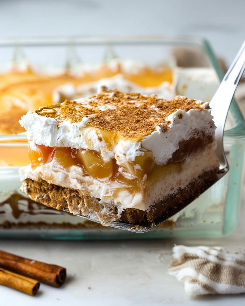 A close-up of a square slice of dessert being lifted from a clear glass dish with a silver spatula. The dessert has four distinct layers: the bottom layer is a dark brown crumbly crust, above it is a creamy light beige layer, then a thick shiny layer of golden apple filling with visible apple pieces, and on top is a fluffy white whipped cream layer sprinkled with light brown cinnamon powder. The dessert and spatula sit on a white marbled surface with two cinnamon sticks nearby. Photo taken with an iphone --ar 4:5 --v 7