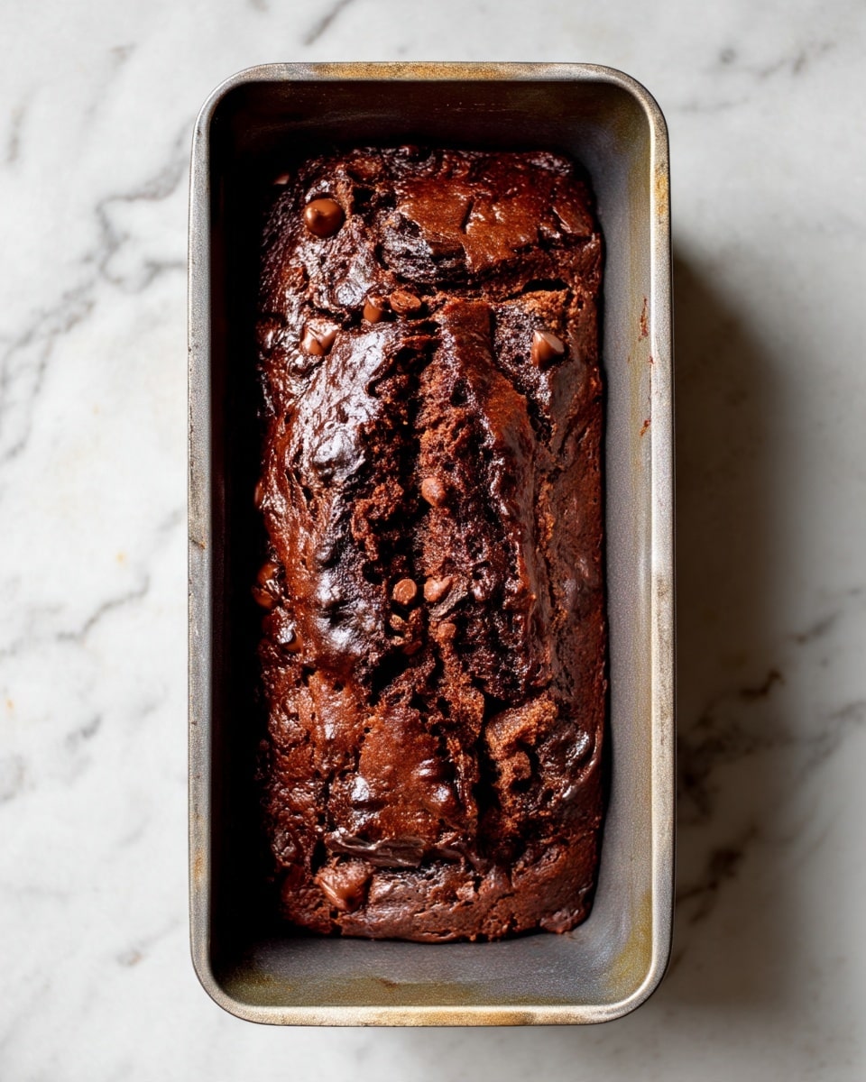A dark chocolate loaf cake sits in a metal baking pan, rising with a cracked top that reveals moist and dense texture. The cake is rich brown with small chunks of chocolate chips embedded throughout, giving it a slightly bumpy surface. The pan has slight wear marks and rests on a white marbled surface that contrasts with the deep color of the cake, showing its glossy, fudgy finish. photo taken with an iphone --ar 4:5 --v 7